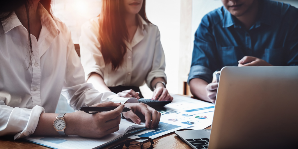 Three people in business attire work together at a table with a laptop, documents, and notebooks. One person writes while the others review charts. Sunlight filters through a window, illuminating the workspace.