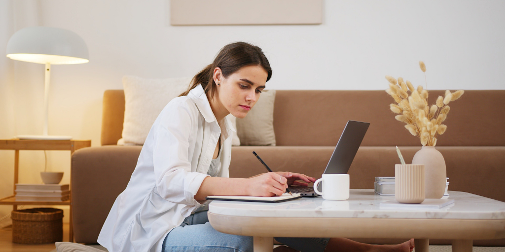 Woman sitting on the floor and working on a laptop on a coffee table. She is writing in a notebook, with a cup beside her. The room has a modern, minimalistic décor with a lamp and dried flowers.