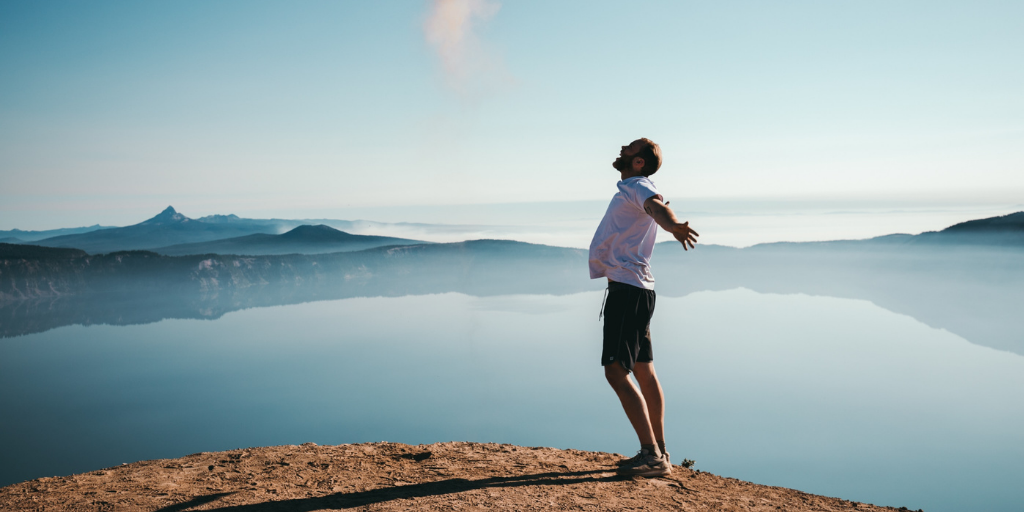 A person stands on a cliff edge with arms outstretched, facing a serene lake reflecting the clear sky. Mountain ranges and a gentle mist form the backdrop under soft, natural light, suggesting a sense of freedom and tranquility.
