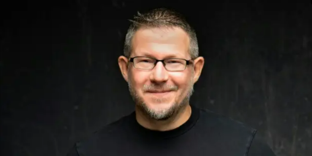 A smiling person with short hair, glasses, and facial hair is wearing a black T-shirt, contemplating whats next for work in 2025 against a plain, dark background.