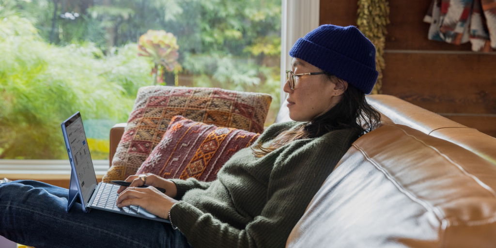 A person wearing a blue beanie and glasses is lying on a couch, using a small laptop. They are dressed in a green sweater and jeans. There are colorful pillows behind them, and a large window shows greenery outside.