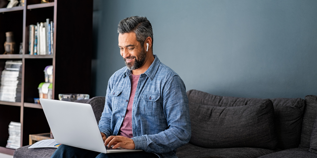 A man with gray hair and a beard, wearing earphones and a casual blue jacket over a maroon shirt, is smiling while using a laptop on a dark gray sofa. A bookshelf with books and a small plant is in the background against a dark wall.