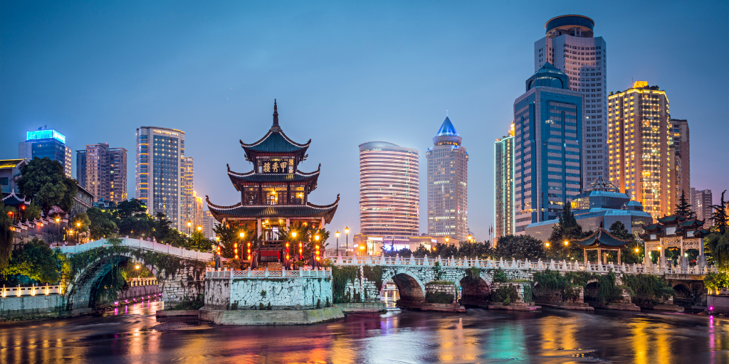 A serene cityscape at dusk featuring a traditional Chinese pavilion illuminated by lights, surrounded by modern skyscrapers. An arched stone bridge crosses a calm river, reflecting the vibrant city lights.