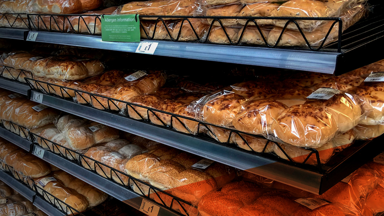 Shelves filled with various packaged bread loaves in a store. The breads are wrapped in clear plastic, each with a price label of £1. A green allergy information sign is visible on the top shelf.