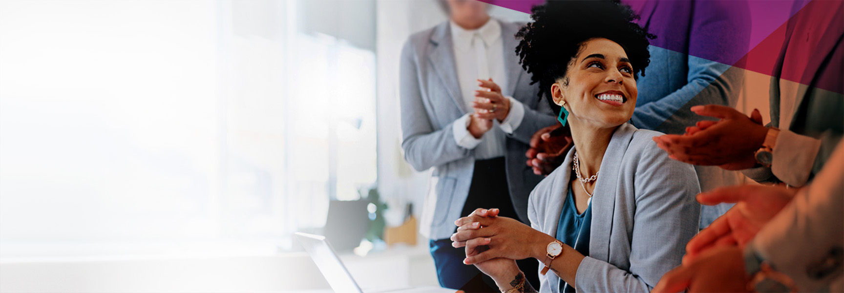 A woman with curly hair sits at a table, smiling and looking to the side. She is wearing a gray blazer and blue top. Several people around her are clapping. The background is bright with a modern office setting.