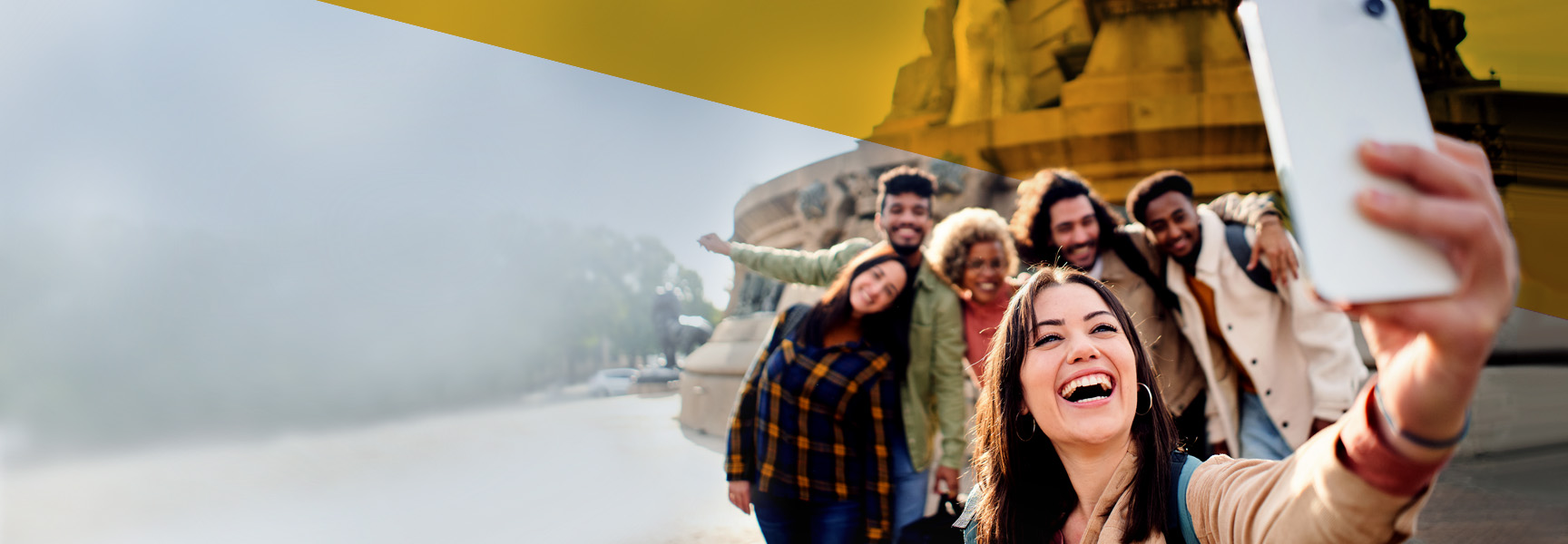 A group of people stand close together, smiling as they take a selfie. The background features a blurred cityscape, suggesting an urban area. The sky above is partly cloudy, and the mood is joyful and lively.