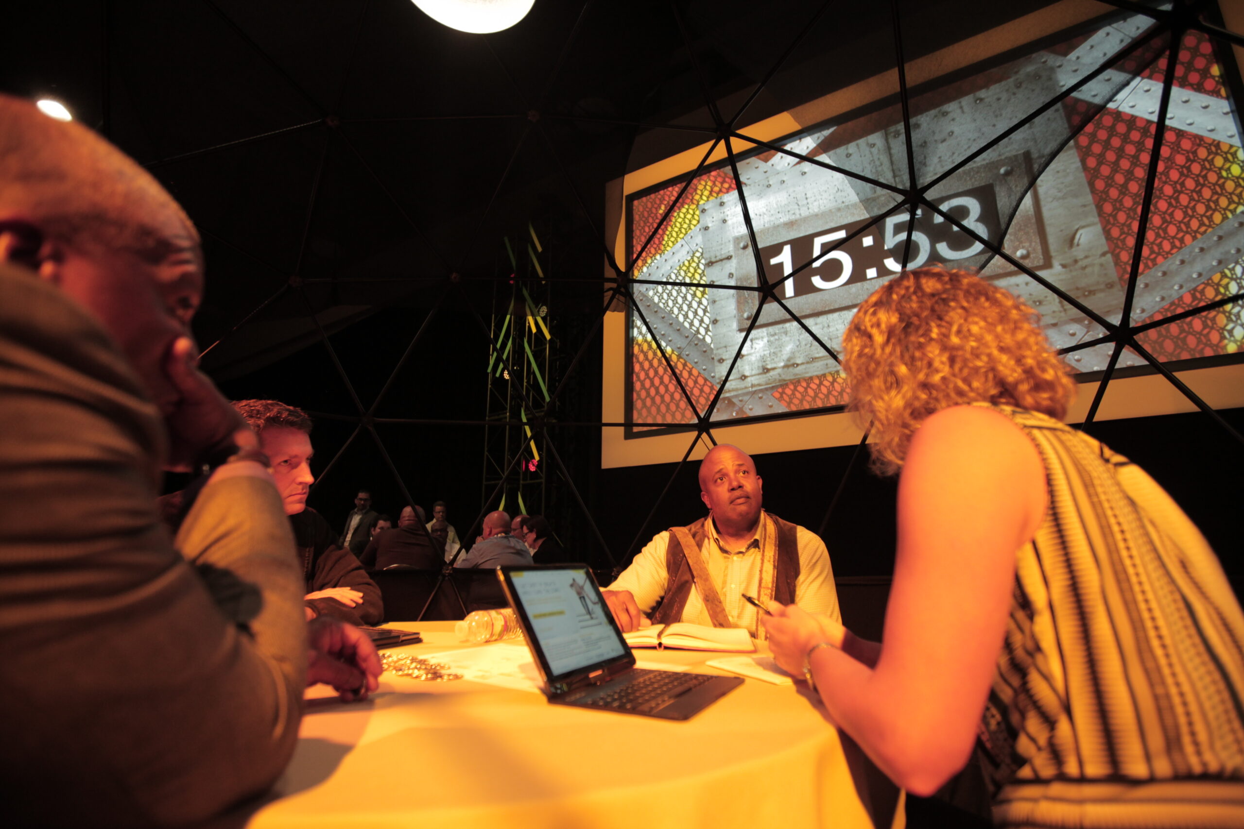 People sit at a round table, engaged in discussion, with a laptop open. A large screen in the background displays 15:53 in white numbers on a colorful geometric pattern. The setting appears to be a dimly lit room with a modern design.