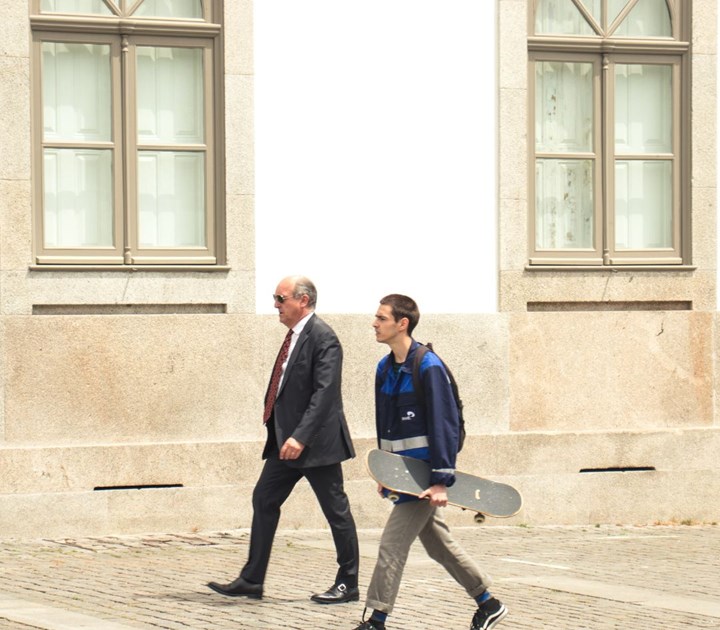 A man in a suit and sunglasses walks beside a young man in a blue jacket holding a skateboard, discussing the nuances of reward strategy as they stroll along the cobblestone path in front of a building with large windows.