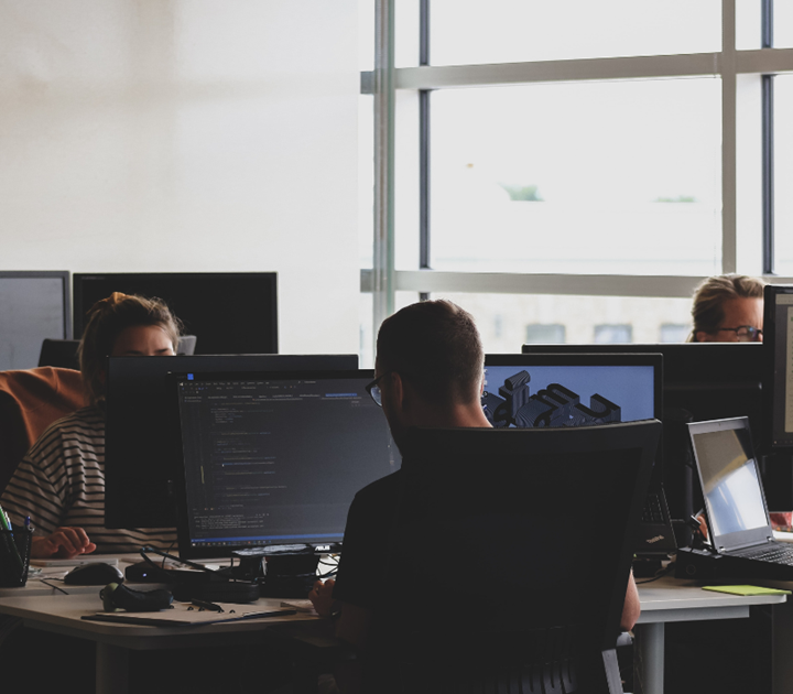 People working in a modern office, concentrating on computer screens. A large window provides natural light. Monitors display coding and design software. The atmosphere seems focused and tech-oriented.