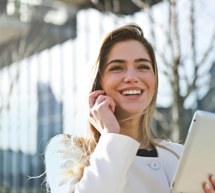 A woman with long hair smiles while holding a tablet and talking on a smartphone. She is wearing a light-colored coat, standing outdoors, with glass buildings and trees in the background.