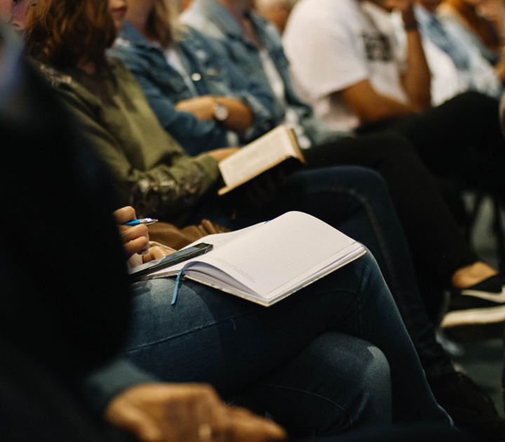People seated closely in a row are focused on an event. Some are holding notebooks and pens, while others hold books. They are dressed casually, and their faces are not visible.