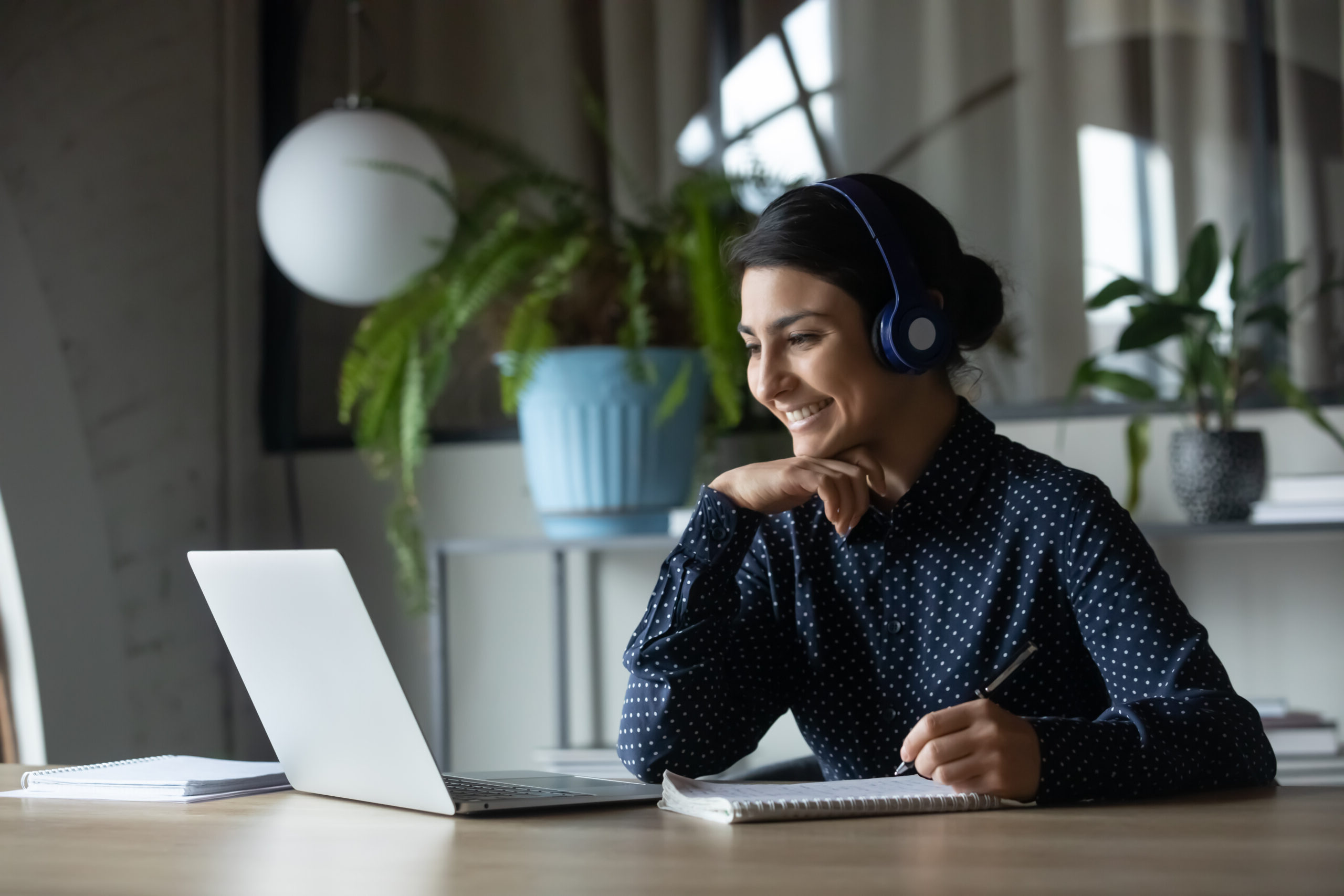 A person wearing headphones sits at a desk, smiling at a laptop displaying appreciation quotes. They have a notebook and pen ready to jot down thoughts. A potted plant and round light fixture add charm to the background.