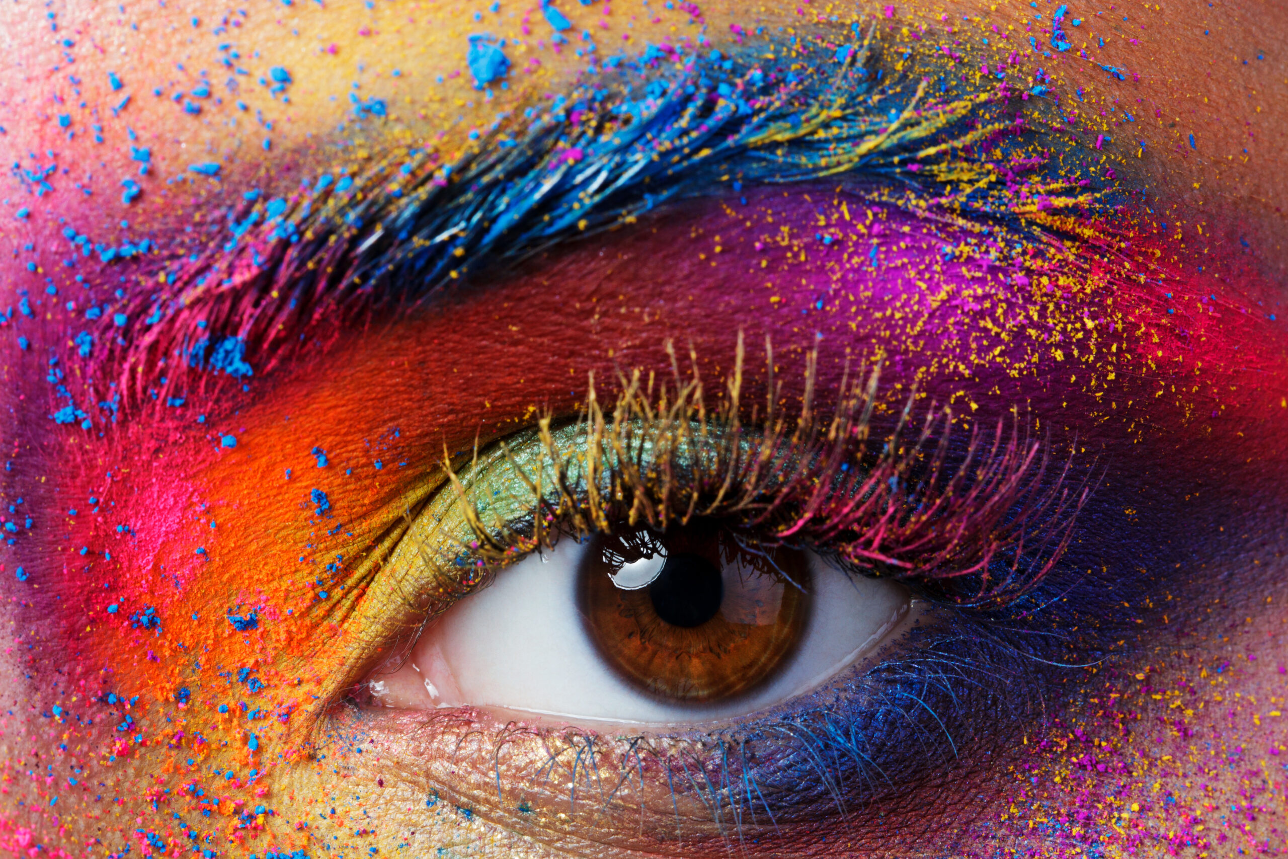Close-up of a brown eye with vibrant, multicolored eyeshadow and powder, including blue, purple, yellow, and pink. The eyebrow is heavily colored, creating a bold and artistic look.