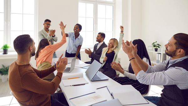 employees in the boardroom with laptops and paper cheering and hi 5ing each other