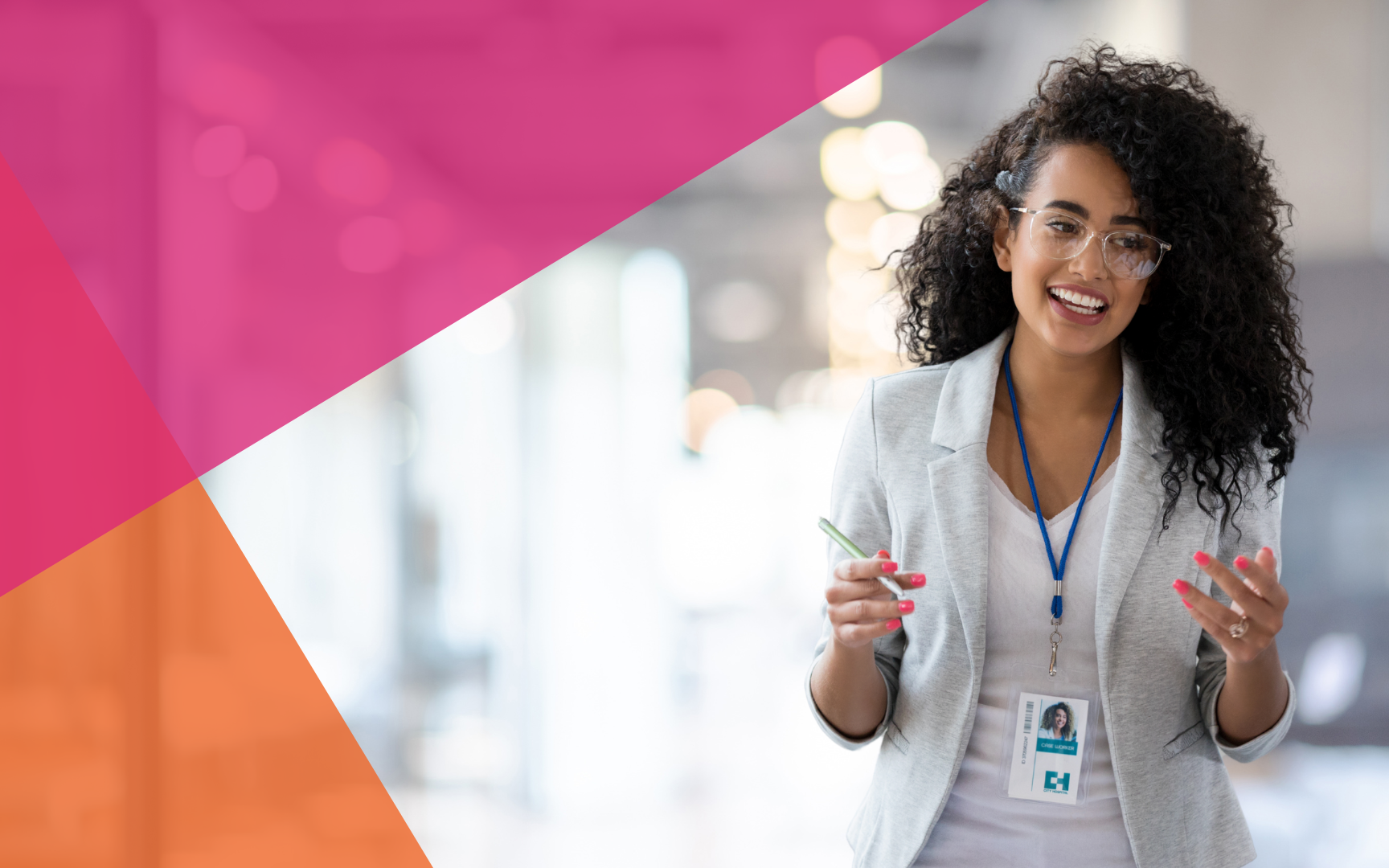 Smiling woman with curly hair wearing glasses, a gray blazer, and a lanyard stands in a modern office. She holds a pen and gestures with her hands. Pink and orange geometric shapes overlay the image corners.