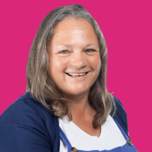 A smiling woman with long, grayish-brown hair wears a white shirt, blue apron, and navy cardigan, posing in front of a solid bright pink background.