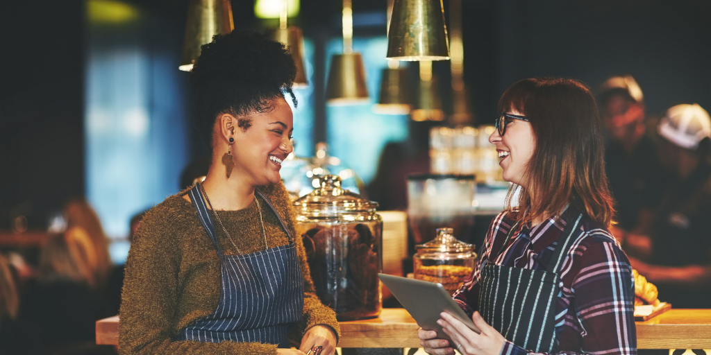 Two women wearing aprons are smiling and talking in a cozy café. One holds a tablet, while the other stands nearby. Large jars and hanging lights are visible on the counter in the background.