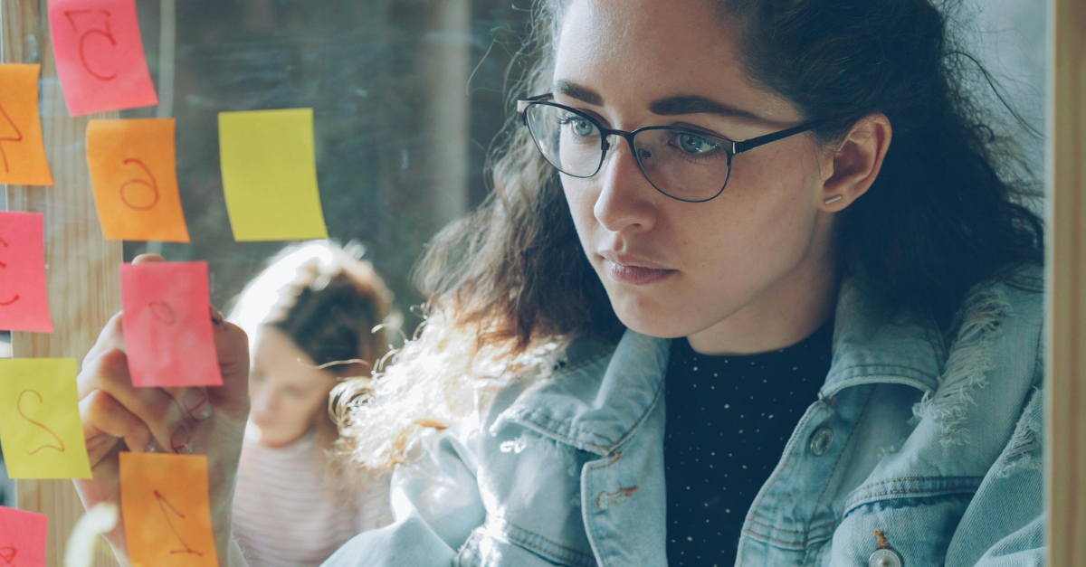 A woman wearing glasses and a denim jacket studies colorful sticky notes on a glass wall, appearing focused. Another person is blurred in the background.