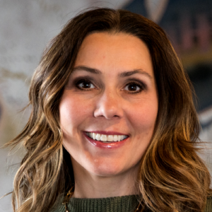 A woman with long wavy brown hair smiles at the camera, wearing a green sweater and a necklace. The background is softly blurred with neutral colors.