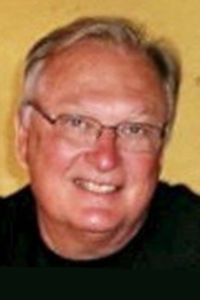 A smiling older man with short gray hair and glasses, wearing a black shirt, poses in front of a yellow background.