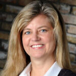 A woman with long blonde hair smiles at the camera. She is wearing a white collared shirt and a dark sweater, standing in front of a brick wall background.