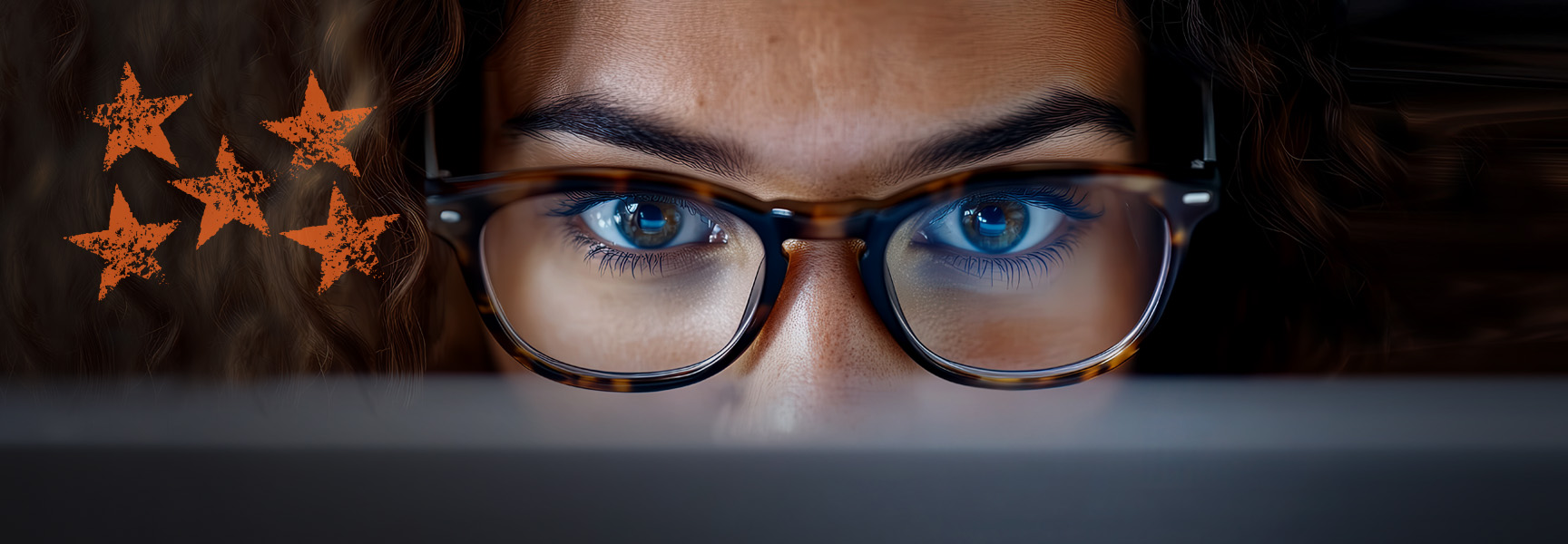 A person with curly hair and glasses intently looks at a laptop screen, with a group of five orange stars drawn on the left side of the image.