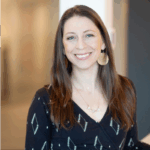 A woman with long brown hair, wearing a dark patterned blouse and large round earrings, smiles while standing indoors in a softly lit space.