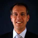 A man in a suit and tie smiles at the camera against a dark background.
