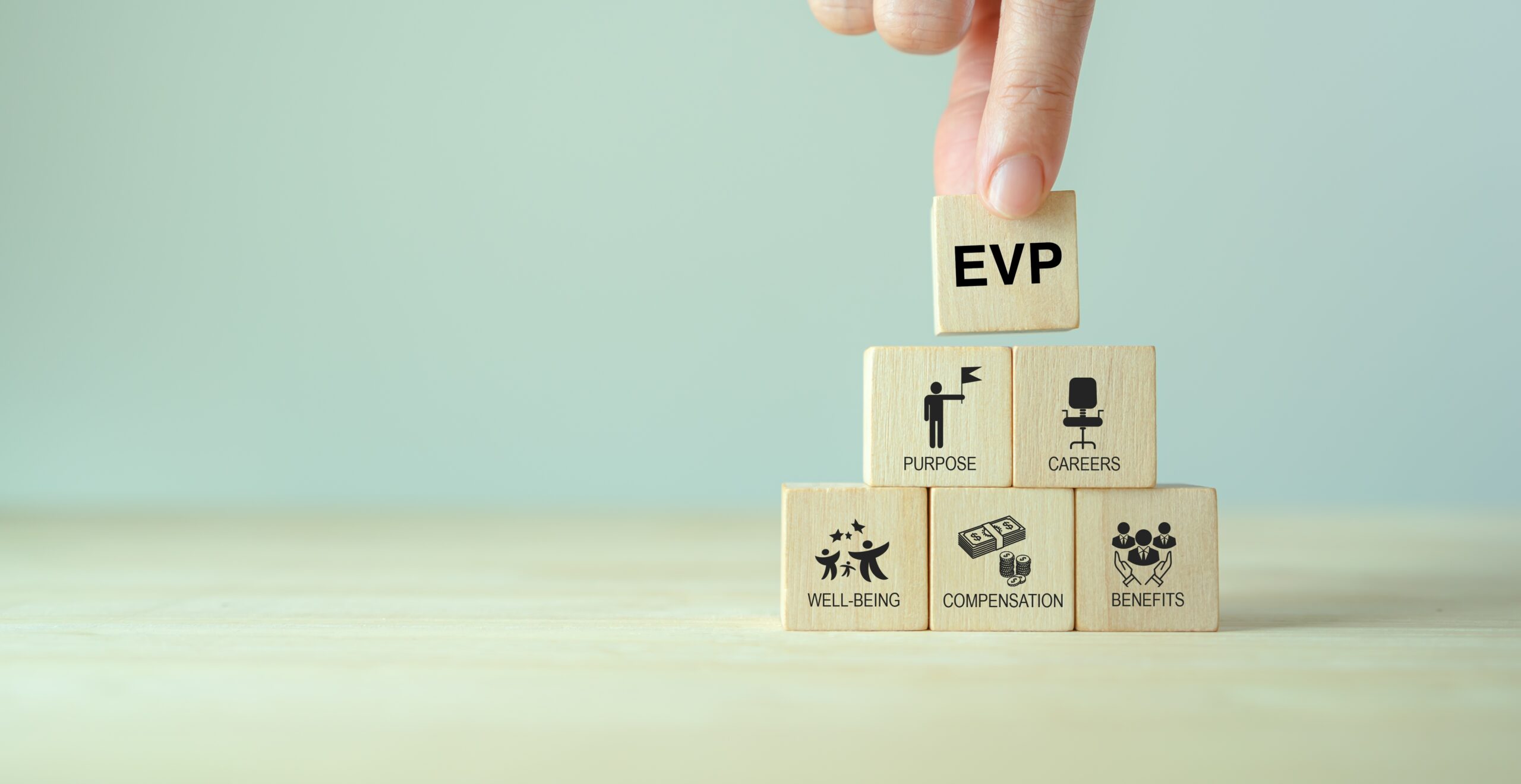 A hand places a block labeled EVP on top of a pyramid of wooden blocks. Below, blocks with icons read: PURPOSE, CAREERS, WELL-BEING, COMPENSATION, and BENEFITS. The background is a pale blue-gray.