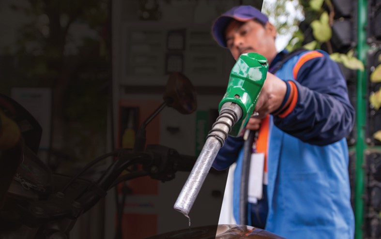A person in a blue uniform refuels a motorcycle with a green gasoline nozzle at a gas station. The background is slightly blurred, highlighting the refueling action.