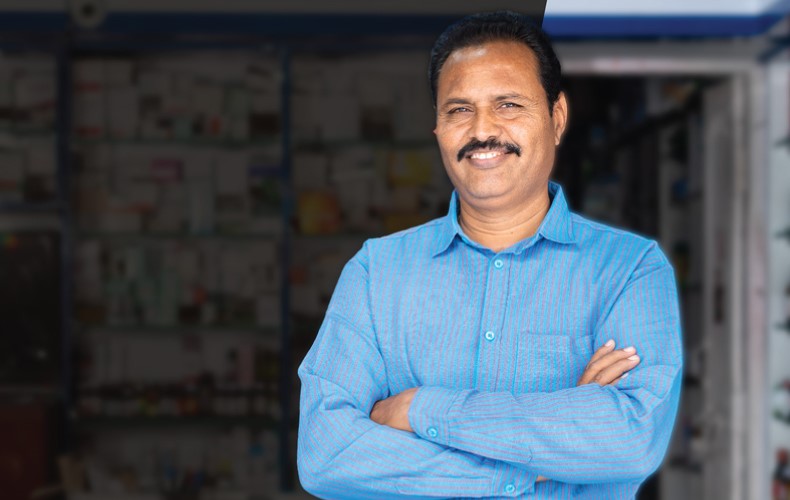 A man in a blue shirt stands with his arms crossed, smiling in front of shelves filled with pharmacy items.