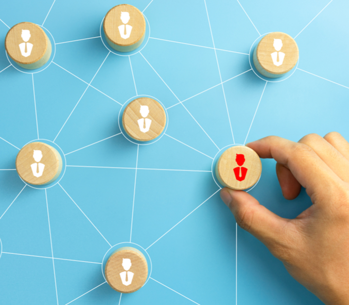 A hand is placing a wooden disc with a red silhouette of a person onto a blue background. The board shows interconnected white lines with other discs featuring white silhouettes, symbolizing a network or team structure.