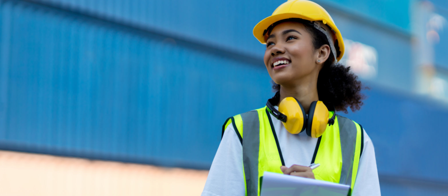A woman wearing a yellow hard hat, yellow headphones around her neck, and a neon safety vest smiles while holding a clipboard. The background shows a blurred blue and orange industrial setting.