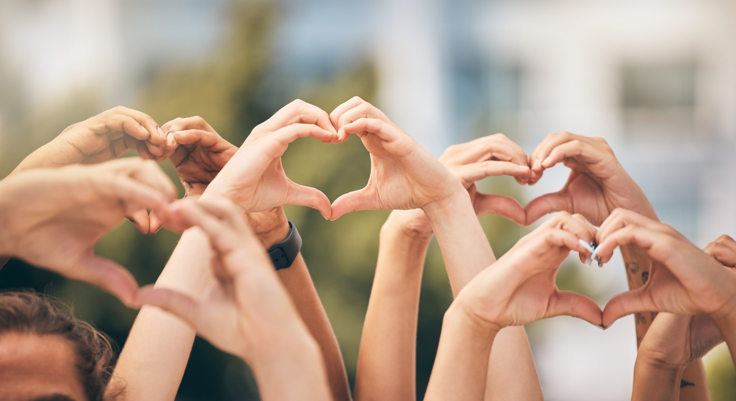 A group of people holding up their hands against a blurred outdoor backdrop, forming heart shapes with their fingers. The focus is on the hands, symbolizing unity and love.
