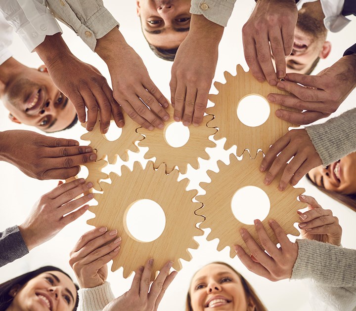 A diverse group of people stands in a circle, each holding wooden gears overhead. The gears interlock at the center, symbolizing unity and teamwork. The people are smiling and facing inward towards the gears.