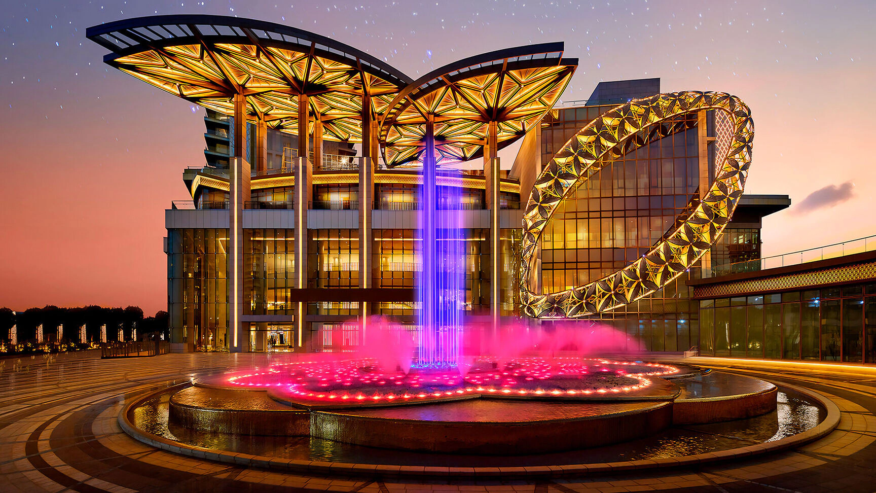 Modern building with a unique geometric roof and a large circular sculpture. A colorful fountain with purple and pink lights is in the foreground, set against a sunset sky.