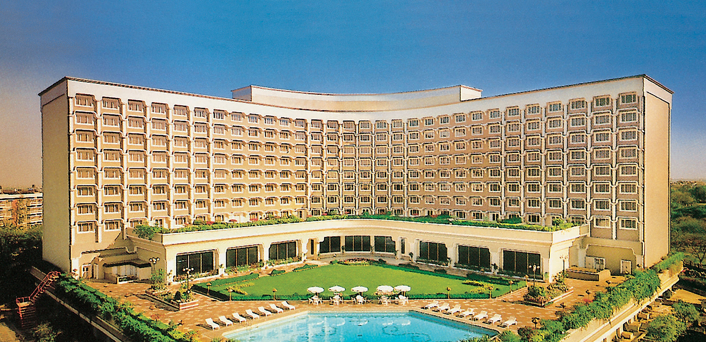 A large, curved hotel building with many windows, surrounded by greenery. In front, there is a swimming pool with lounge chairs and umbrellas on a sunny day. The sky is clear and blue.