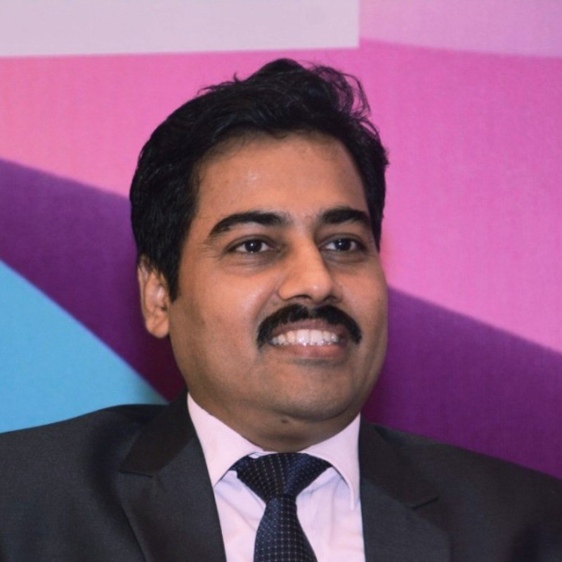 A man in a dark suit and tie smiles while seated in front of a colorful, abstract background with shades of pink, purple, and blue.