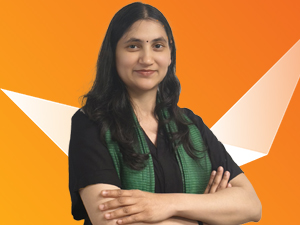 A woman with long dark hair and a green scarf stands confidently with arms crossed, smiling, in front of an orange background with white geometric shapes.