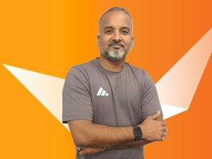 A man with short hair and a beard stands with arms crossed, wearing a gray t-shirt and a black watch, against an orange background with white geometric shapes.