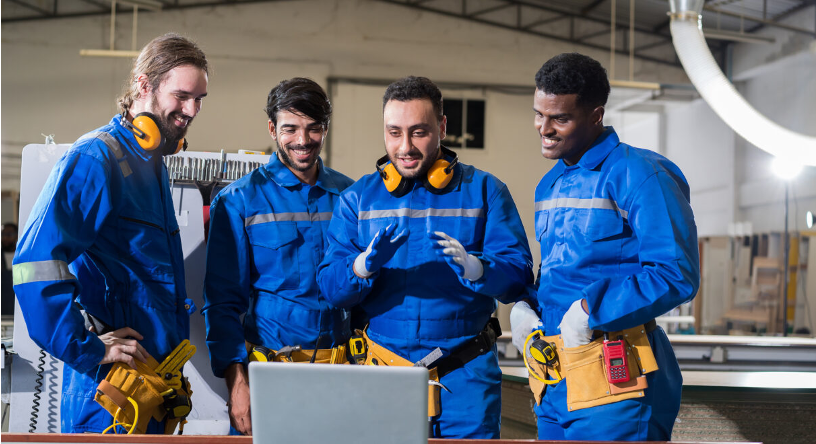 Four male workers in blue coveralls and safety gear smile and look at a laptop screen, appearing engaged and collaborative in an industrial workshop setting.