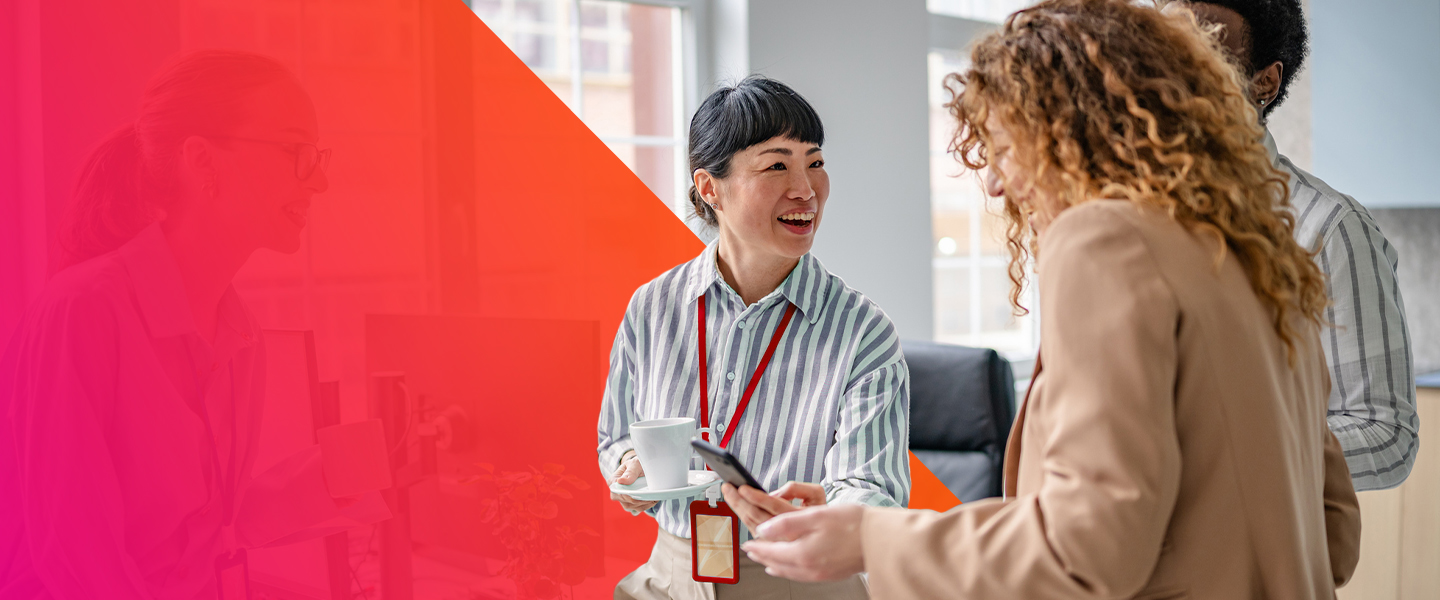 Two women in business attire smile and talk in a bright office while holding coffee cups. One woman also holds a smartphone. A pink and red geometric overlay covers the left side of the image.