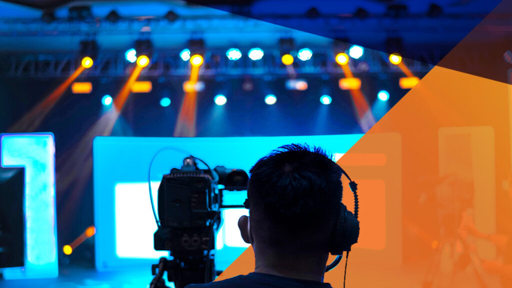 A person wearing headphones operates a video camera, filming a brightly lit stage with blue and orange lights and abstract graphic overlays.
