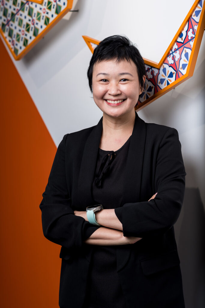 A woman with short black hair, wearing a black blazer and black top, stands smiling with arms crossed. Colorful geometric wall art and orange accents are visible in the background.