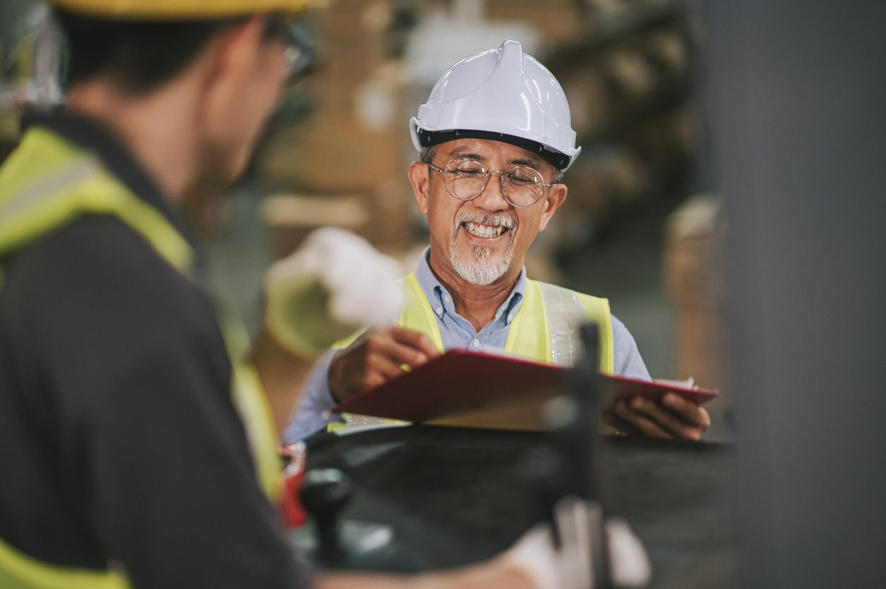 An older man wearing a white safety helmet and yellow vest smiles while writing on a clipboard in an industrial setting, interacting with another worker in the foreground.