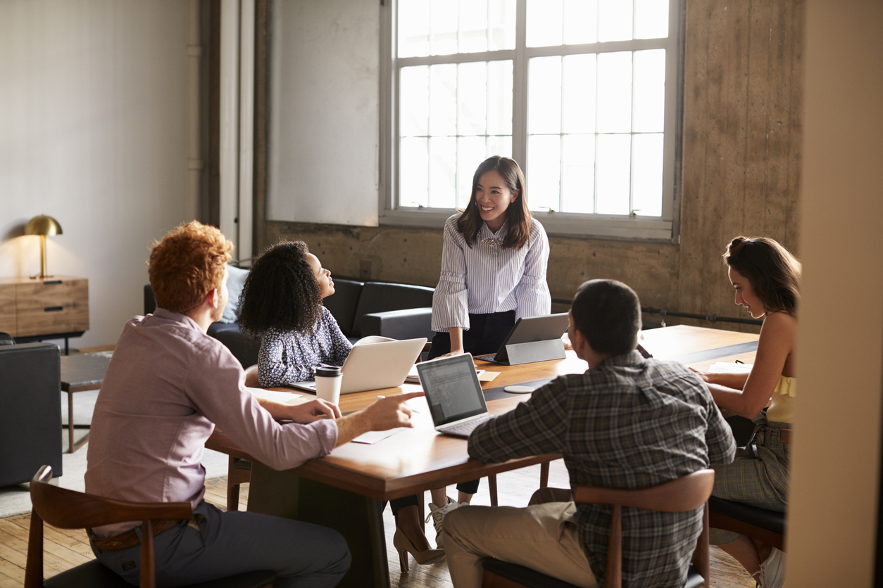 Five people sit around a table in a modern office, having a discussion. One woman stands and smiles while speaking to the group. Laptops and notebooks are on the table, and sunlight streams through a large window.