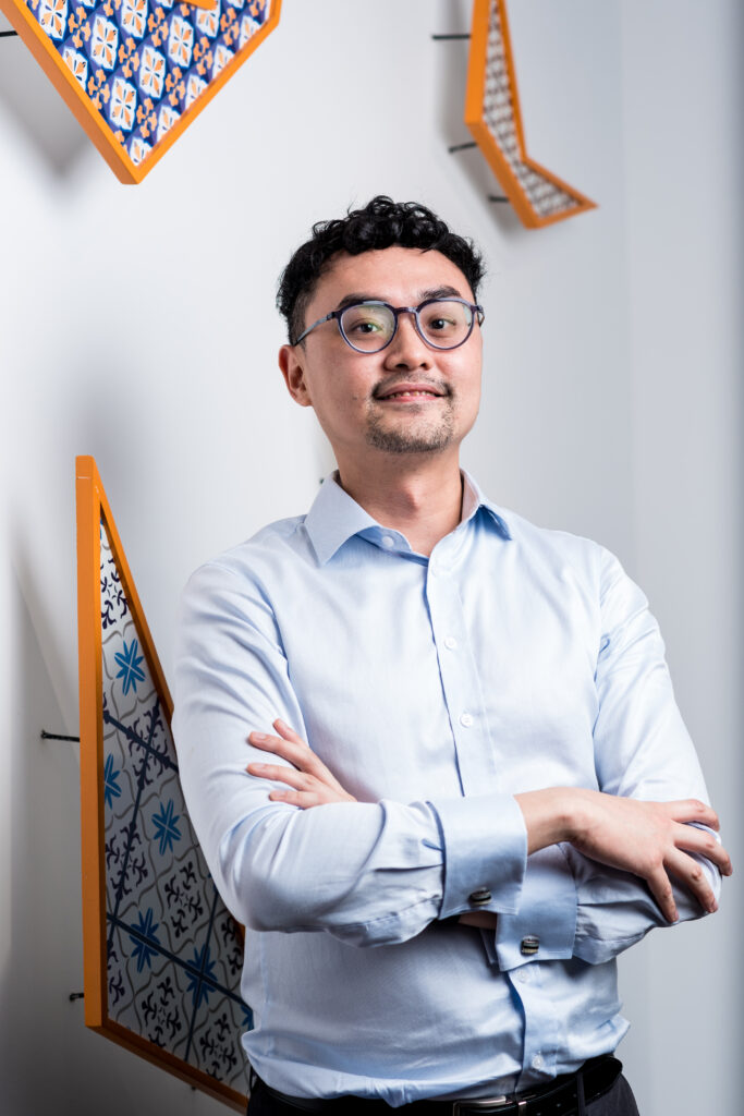 A man with short curly hair, glasses, and a light blue shirt stands with arms crossed, smiling slightly. Colorful, geometric wall art with tile patterns decorates the white wall behind him.