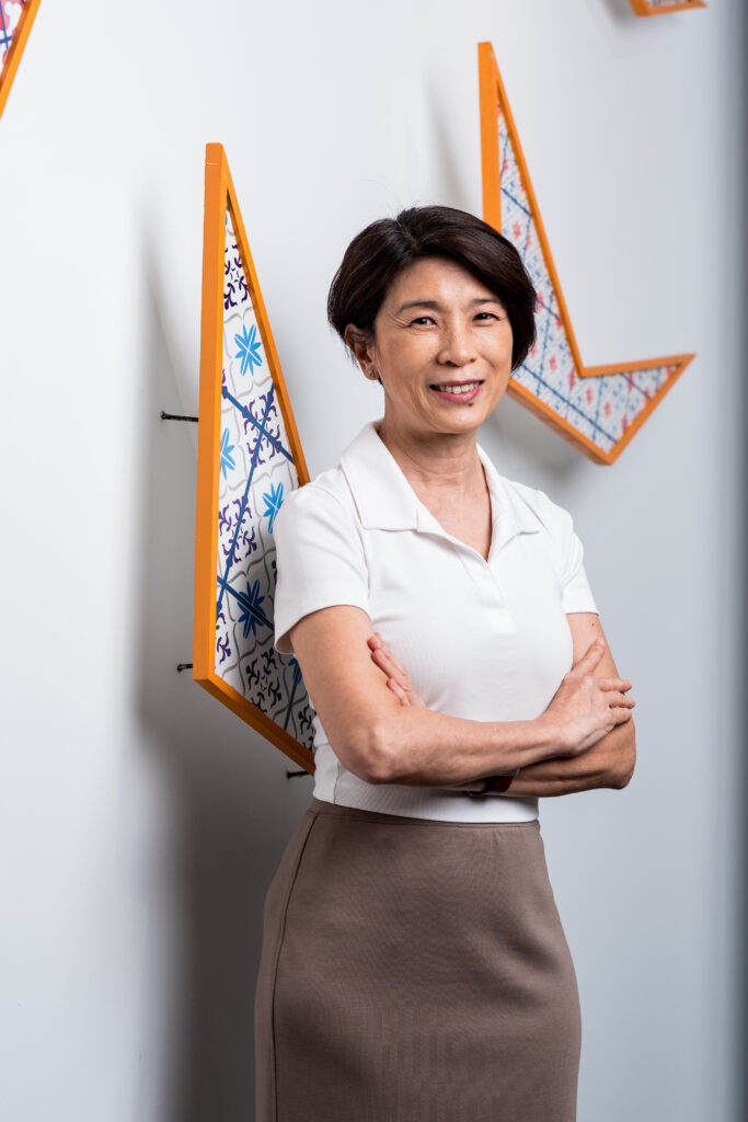 A woman with short dark hair, wearing a white collared shirt and taupe skirt, stands with arms crossed and smiles. Geometric, colorful wall art is displayed behind her on a white wall.