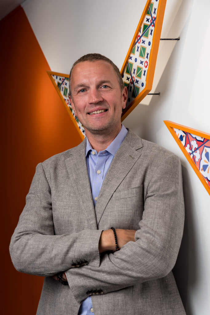 A man in a light gray blazer and blue shirt stands with arms crossed, smiling in front of a white wall with geometric, colorful decorative pieces and an orange accent wall.