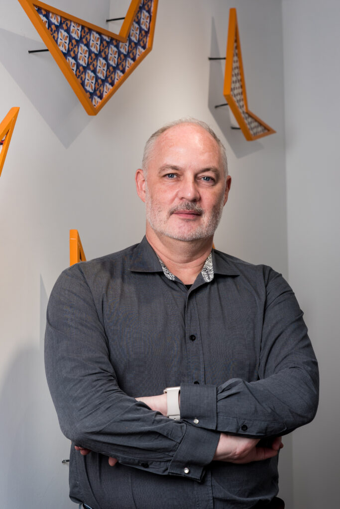 A middle-aged man with short gray hair and a beard stands with arms crossed, wearing a dark gray shirt. Geometric orange and patterned wall art is displayed behind him on a white wall.